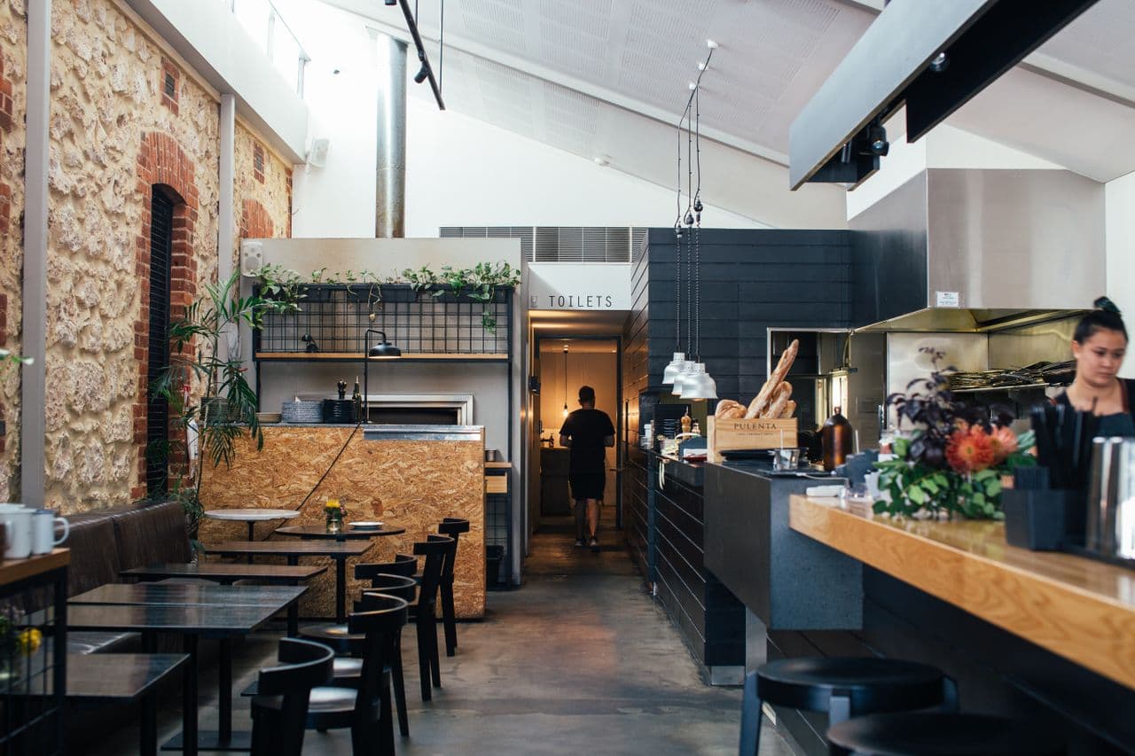 Industrialne wnętrze restauracji z ceglaną ścianą, drewnianymi stolikami i otwartą kuchnią